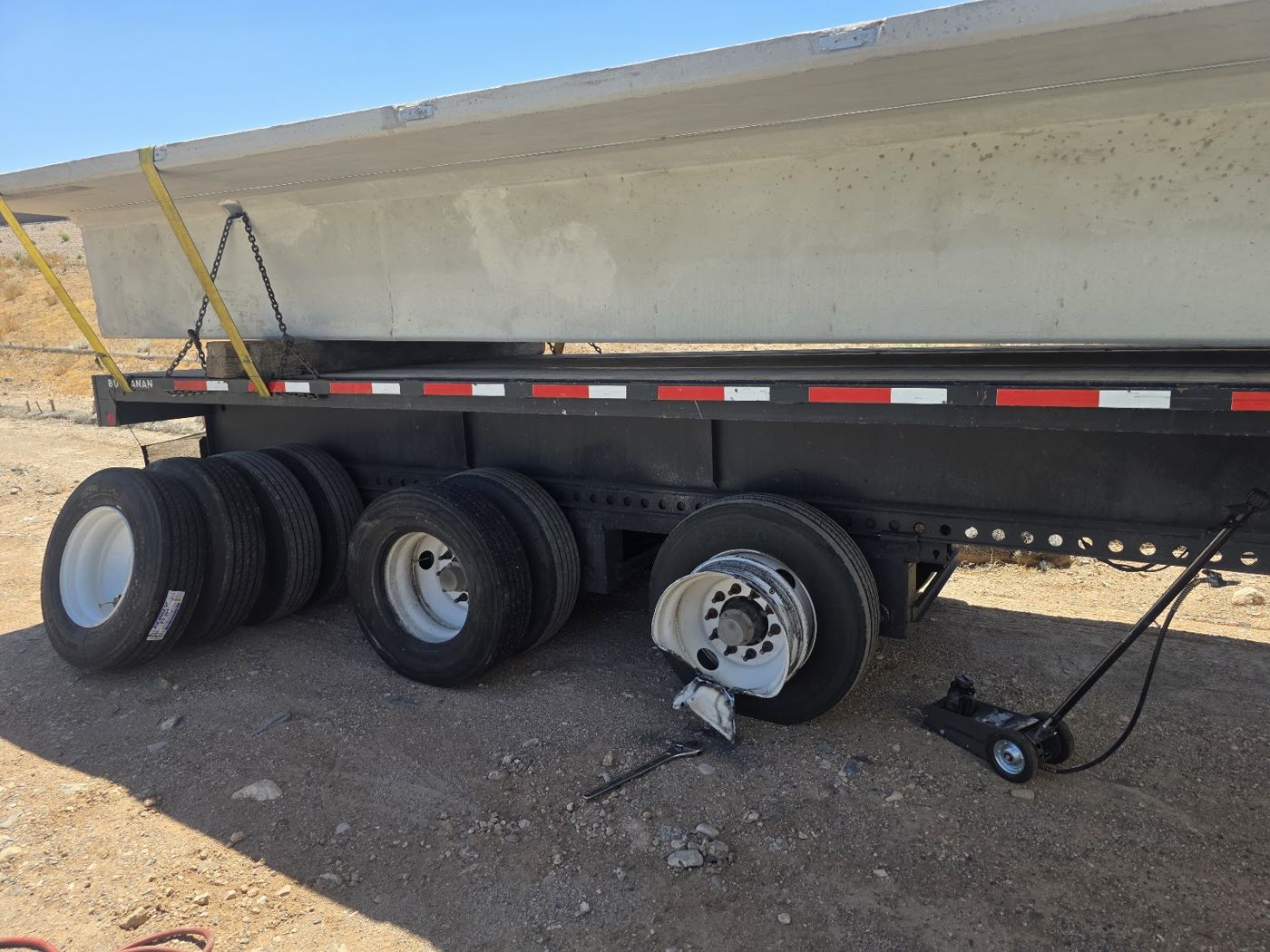 A flatbed trailer with a wheel off and a jack under the axle, spare tires stacked beside it